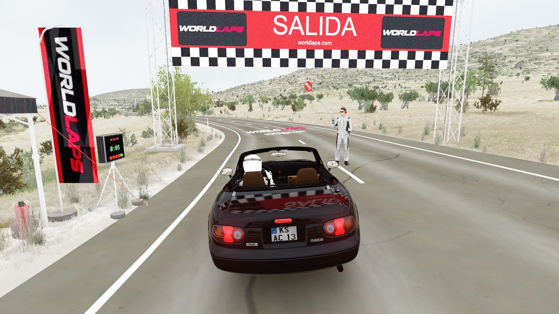 Captura de Assetto Corsa en la salida del tramo de Los Herreros: el coche bajo el pórtico de SALIDA en una curva a la izquierda, con olivos y lomas al fondo.